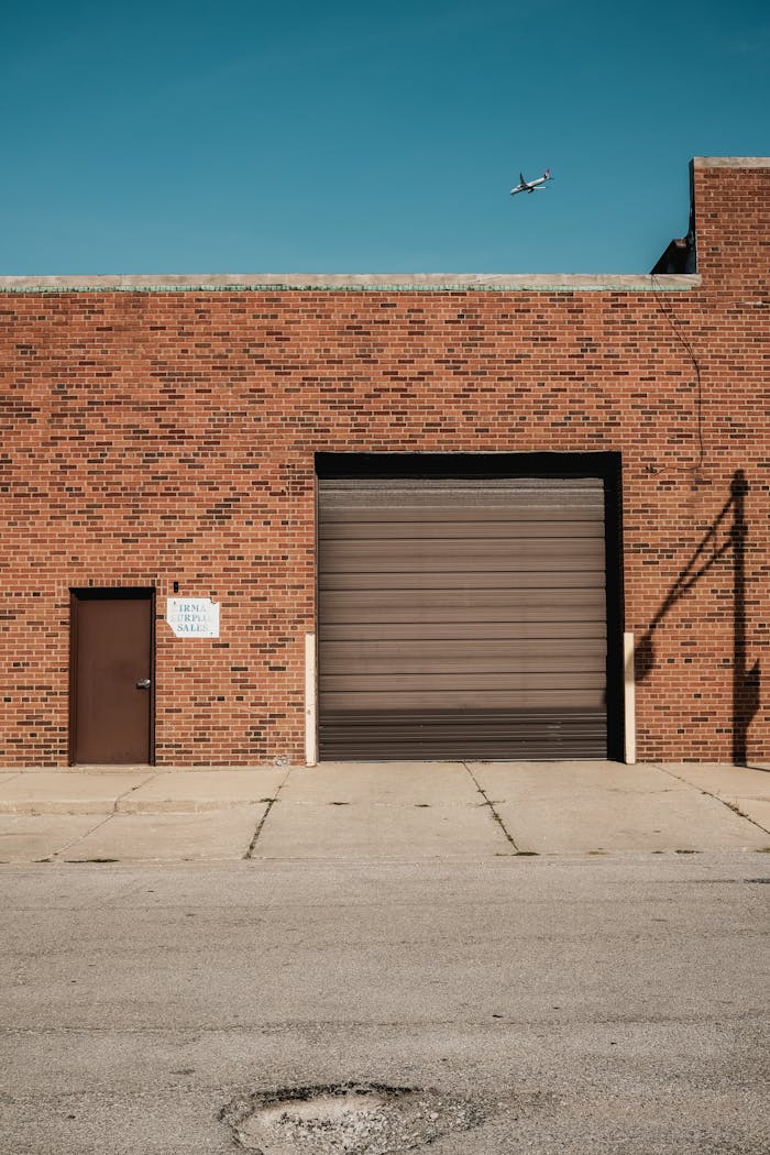 About A brick industrial building with a garage door and airplane in the clear sky above.